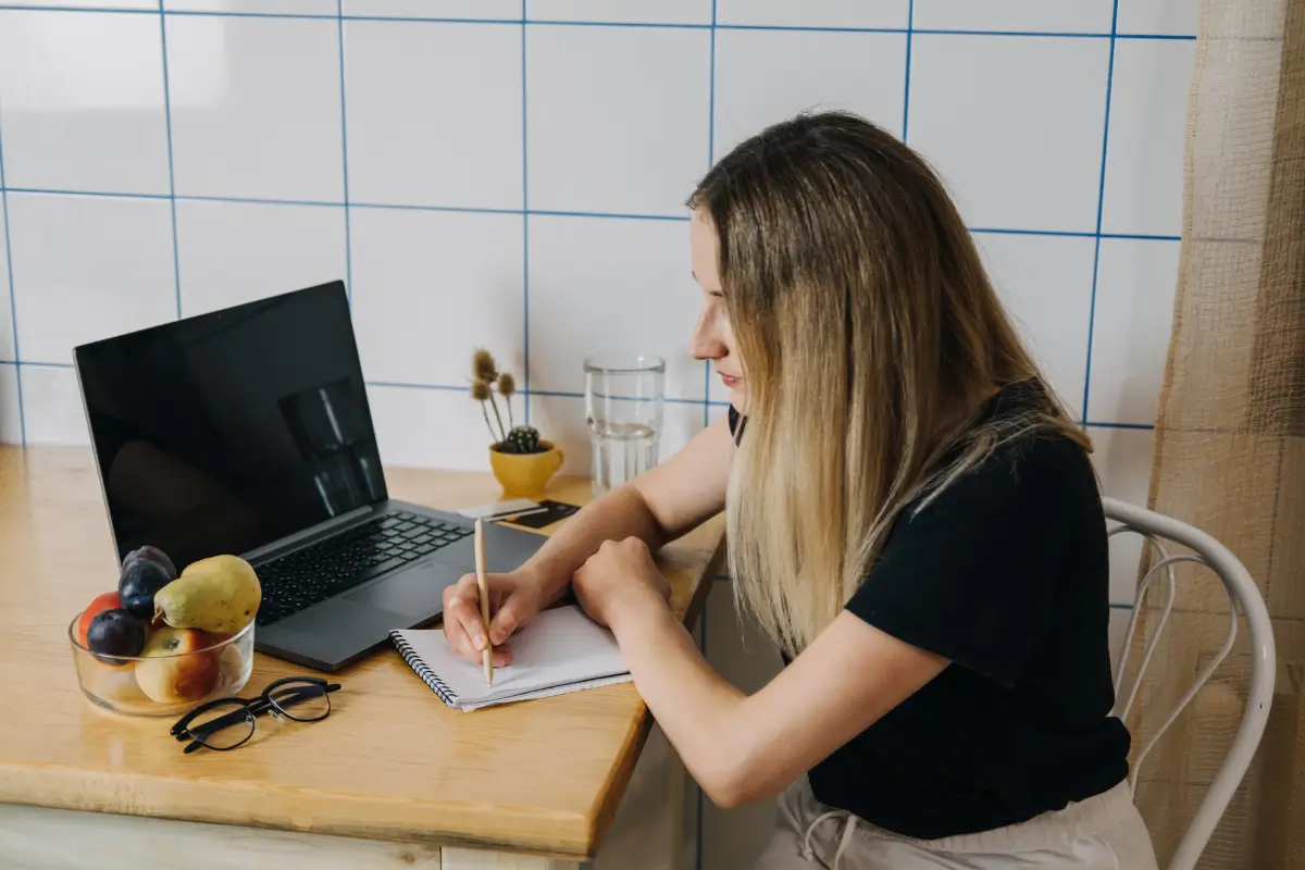 Ruhiger Moment am Küchentisch: Eine Frau schreibt ihr Haushaltsinventar, Laptop und Notizen griffbereit. Praktische Vorbereitung mit System für den Notfallplan.