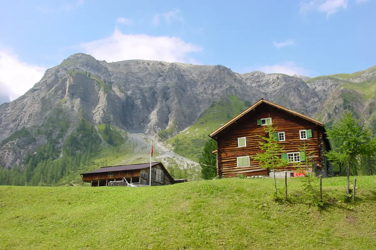 Ein traditionelles Holzhaus steht allein auf einer grünen Bergwiese vor steiler Felskulisse. Sinnbild für abgelegene Lebensweise, Selbstversorgung und ländliche Krisenvorsorge.